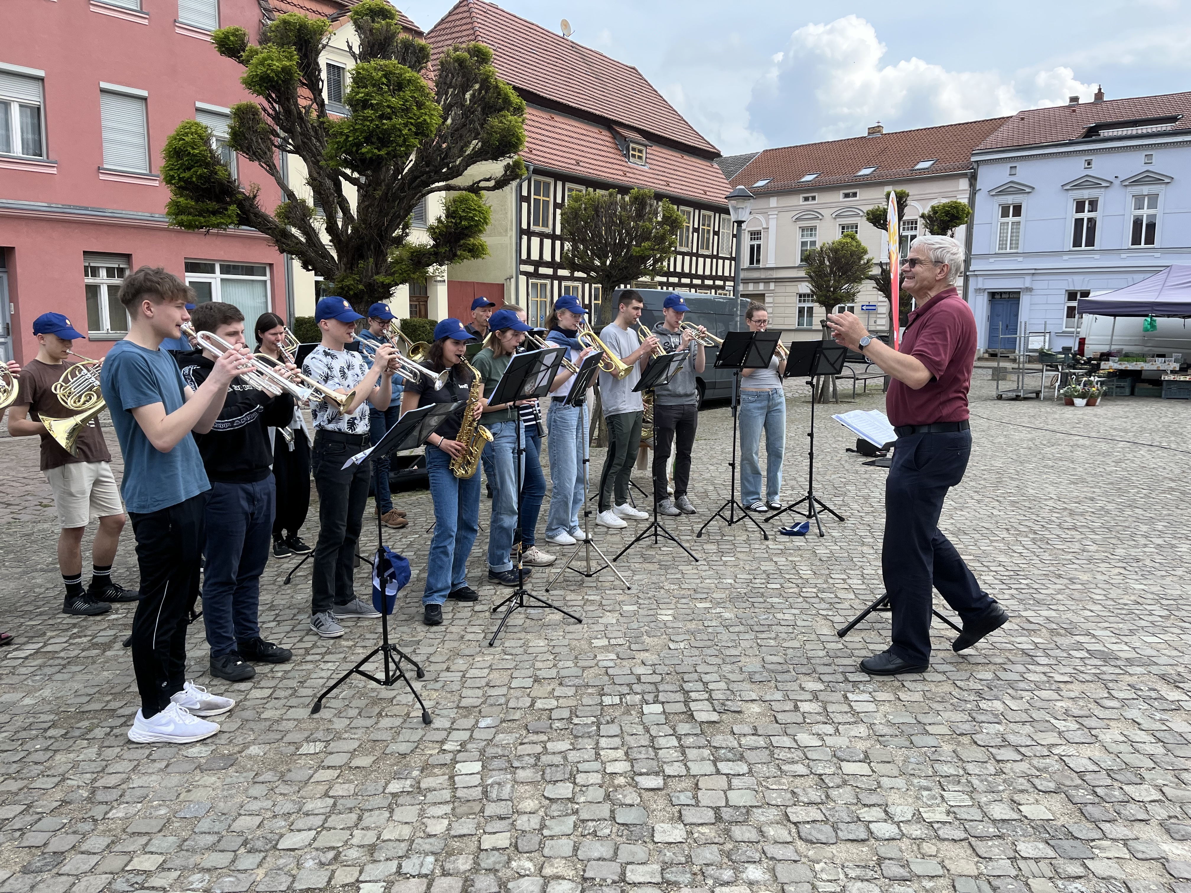 Eine Gruppe junger Blasmusiker spielt in der Öffentlichkeit. Die Gruppe wird von einem Dirigent angeleitet. 