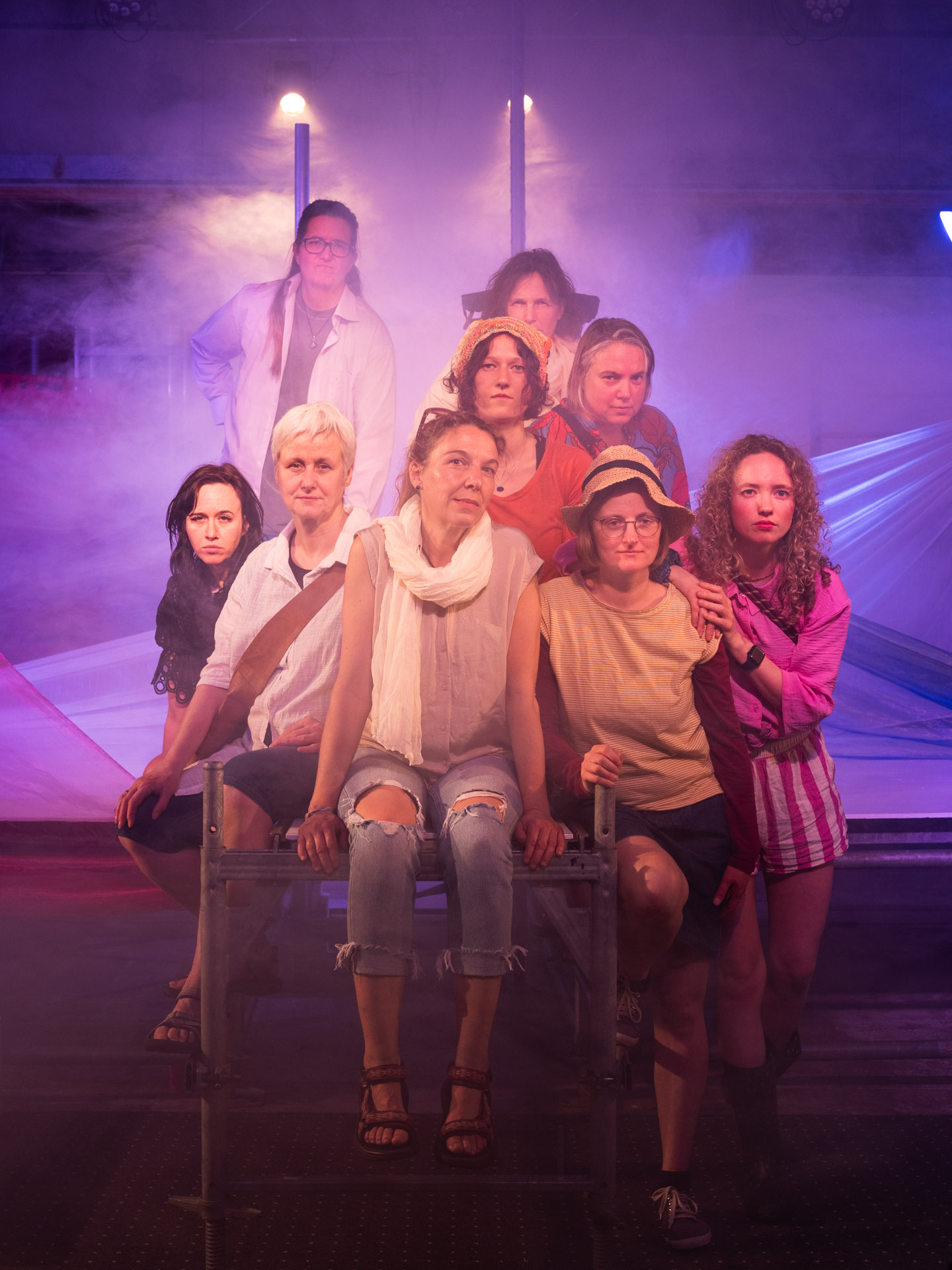 Gruppenfoto von acht Frauen auf einer nebligen Theaterbühne, die in atmosphärisches violettes und rötliches Licht getaucht ist. Drei der Frauen sitzen im Vordergrund auf einem Metallgerüst, während die anderen dahinter posieren. Der Blick aller Frauen ist ernst und direkt in die Kamera gerichtet.