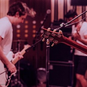 Eine Musikgruppe in einem rötlich ausgeleuchteten Proberaum. Im Vordergrund ist der Hals einer E-Gitarre zu sehen, im Hintergrund spielen zwei Musiker.